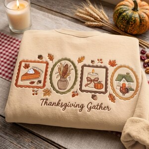 Pode incluir: Uma camisola bege com bordados de uma fatia de tarte de abóbora, uma espiga de milho num vaso, uma vela com uma abóbora e uma casa com um peru, emoldurados. O texto "Thanksgiving Gather" está bordado na parte inferior.