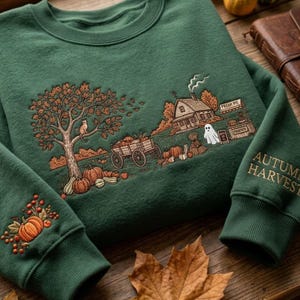 Pode incluir: Uma camisola verde-escura com uma cena de outono bordada, apresentando uma árvore, um gato, uma carroça de madeira cheia de abóboras, uma casa, um fantasma e uma banca de mercado. O texto AUTUMN HARVEST está bordado na manga com motivos de abóboras.
