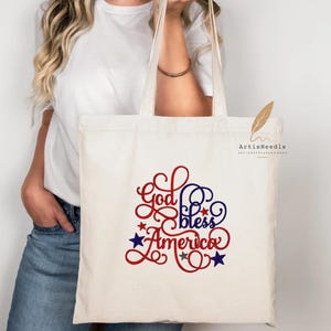 May include: A cream-colored canvas tote bag featuring the text God Bless America in a decorative red and blue script font. The design includes small red, blue, and grey stars scattered around the lettering, creating a patriotic visual theme.
