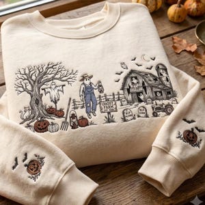 Könnte beinhalten: Ein cremefarbenes Sweatshirt mit Rundhalsausschnitt und schwarzen Halloween-Stickereien. Das Motiv zeigt einen Geist, eine Scheune mit der Aufschrift Ghost Farm, eine Vogelscheuche, Kürbisse, Grabsteine und einen Traktor. An jedem Ärmel ist ein Kürbis.