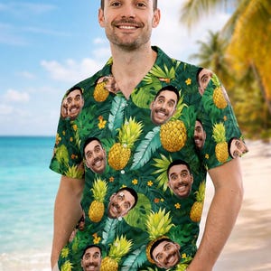 Könnte beinhalten: Grünes Hawaiihemd mit einem sich wiederholenden Muster aus Ananas und Gesichtern. Das kurzärmelige Hemd hat ein tropisches Design mit gelben Blumen und großen grünen Blättern. Das Hemd wird von einer Person am Strand getragen.