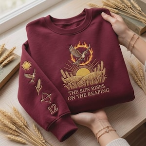 Könnte beinhalten: Ein kastanienbraunes Sweatshirt mit Rundhalsausschnitt und goldener Stickerei eines Vogels in einer Sonne über einem Weizenfeld. Der Text lautet THE SUN RISES ON THE REAPING. Der Ärmel zeigt eine Sonne, einen Vogel, eine Weizenähre, einen Bogen und die 12.
