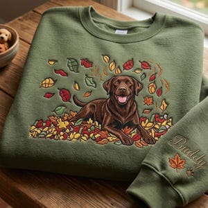 May include: An olive green crewneck sweatshirt featuring an embroidered brown Labrador retriever lying in a pile of colorful autumn leaves. The sleeve is embroidered with the name Buddy, a maple leaf, and a small paw print in matching thread.