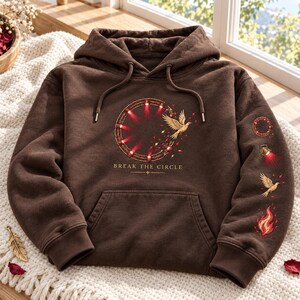 May include: A brown hooded sweatshirt featuring a graphic of a circular structure with red lights, a flying bird, and the text "BREAK THE CIRCLE". The sleeve displays a vertical sequence of a circle, a spotlight, a bird, and a flame graphic.