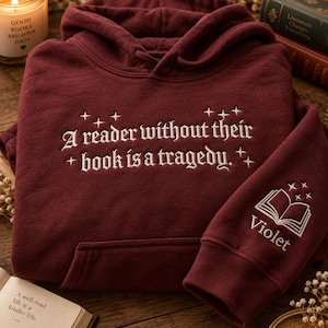 Pode incluir: Uma camisola com capuz em bordeaux escuro com texto bordado a branco que diz A reader without their book is a tragedy. rodeado por pequenas estrelas. A manga apresenta um gráfico bordado de um livro aberto sobre o nome Violet.