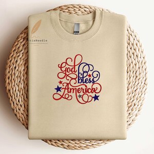 May include: A beige crewneck sweatshirt featuring the embroidered text God bless America in red and blue cursive script. The design includes small red, blue, and grey stars scattered around the lettering. The fabric has a smooth, solid finish.