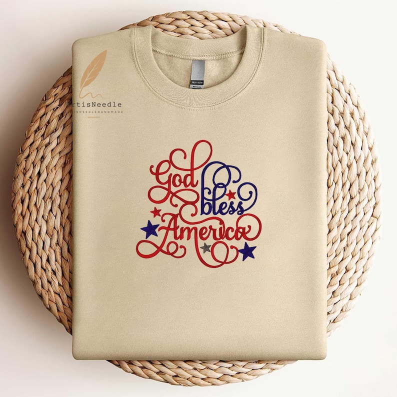 May include: A beige crewneck sweatshirt featuring the embroidered text God bless America in red and blue cursive script. The design includes small red, blue, and grey stars scattered around the lettering. The fabric has a smooth, solid finish.