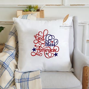 May include: A white square throw pillow featuring the red and blue embroidered text God bless America in a decorative script font. The design is accented with small red, blue, and silver stars scattered around the lettering on the front surface.
