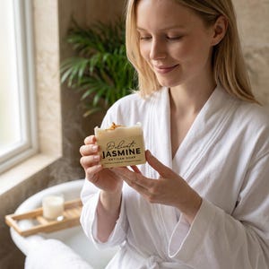 May include: A rectangular bar of off-white artisan soap wrapped in a beige paper label with the text Delicate JASMINE ARTISAN SOAP printed in black and gold. The top of the soap bar is decorated with small, dried yellow flower petals.