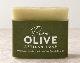 Puede incluir: Una pastilla de jabón rectangular de color verde pálido envuelta en una banda de papel de color verde oliva oscuro. La etiqueta presenta el texto blanco Pure OLIVE ARTISAN SOAP y el texto menor Handmade in Scotland with Unrefined Organic Olive Oil.