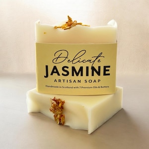 May include: A rectangular bar of off-white soap topped with dried yellow flower petals. A pale yellow paper label wraps around the center, printed with the text Delicate JASMINE ARTISAN SOAP Handmade in Scotland with 7 Premium Oils & Butters.