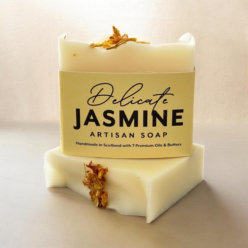 May include: A rectangular bar of off-white soap topped with dried yellow flower petals. A pale yellow paper label wraps around the center, printed with the text Delicate JASMINE ARTISAN SOAP Handmade in Scotland with 7 Premium Oils & Butters.