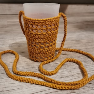 Peut inclure: Un porte-gobelet en crochet avec une longue sangle. Le porte-gobelet est jaune moutarde et présente un motif en crochet. La sangle est faite du même fil et est suffisamment longue pour être portée sur l'épaule.
