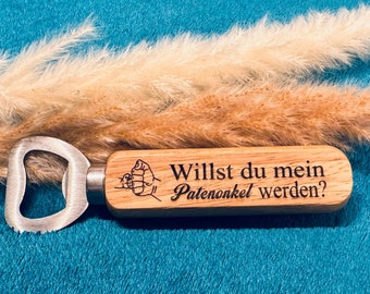 May include: A wooden bottle opener with the text "Willst du mein Patenonkel werden?" is displayed. The opener has a silver metal head and a light brown wooden handle. The background includes dried ornamental grass.