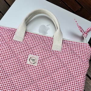 Könnte beinhalten: Rote und weiße Gingham-Laptop-Hülle mit einem weißen Canvas-Griff und einem kleinen weißen Etikett mit dem Text "NUR" in Schwarz.