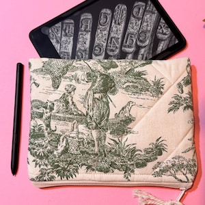 May include: A beige quilted zippered pouch with a green toile pattern featuring a pastoral scene. The pouch has a white rope zipper pull. A black stylus is lying next to the pouch.