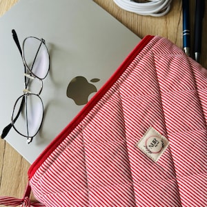 Könnte beinhalten: Eine rot-weiß gestreifte, gepolsterte Laptoptasche mit rotem Reißverschluss und kleinem Etikett. Ein silberner Laptop, eine Brille, Stifte und ein Ladegerät liegen auf einer Holzoberfläche.
