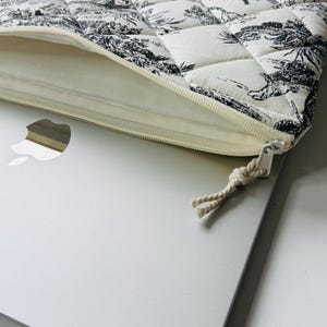 May include: A quilted laptop sleeve featuring a black and white toile pattern. The sleeve has a cream-colored zipper and a matching fabric lining. The design includes a landscape scene. The sleeve is partially open, revealing a silver laptop.
