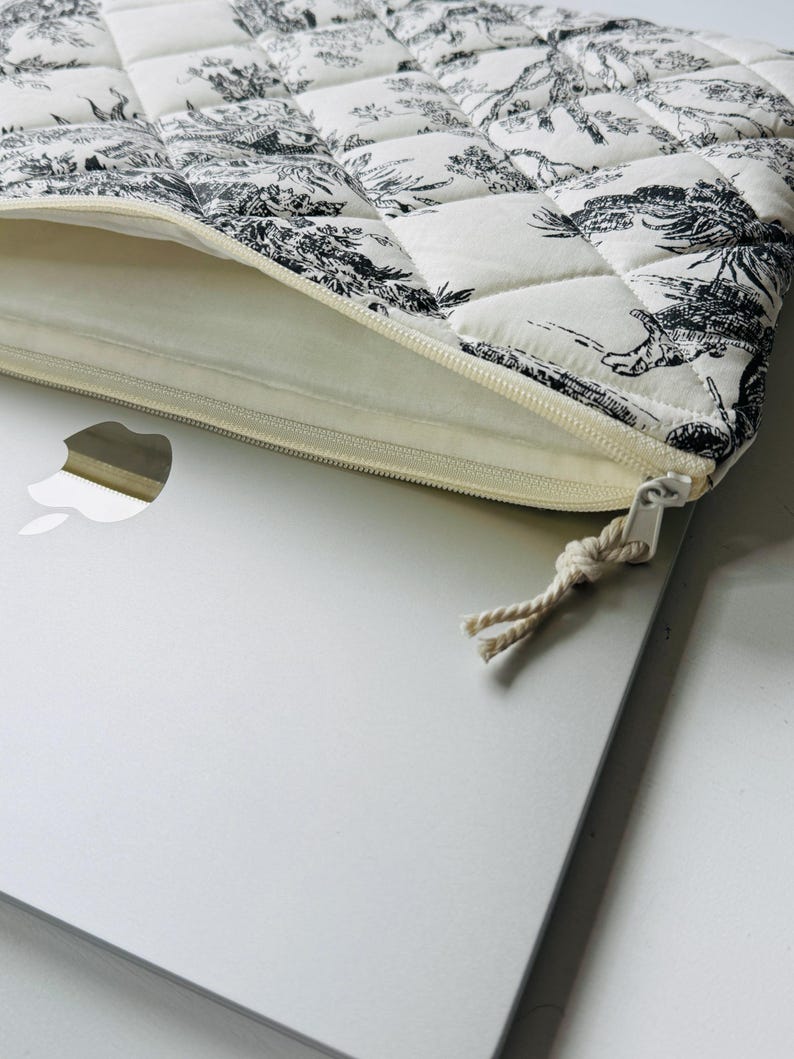May include: A quilted laptop sleeve featuring a black and white toile pattern. The sleeve has a cream-colored zipper and a matching fabric lining. The design includes a landscape scene. The sleeve is partially open, revealing a silver laptop.
