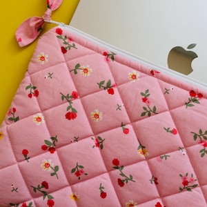 Pode incluir: Capa acolchoada para laptop rosa com fecho de correr e estampado floral. A capa tem uma pequena alça de tecido na parte superior.