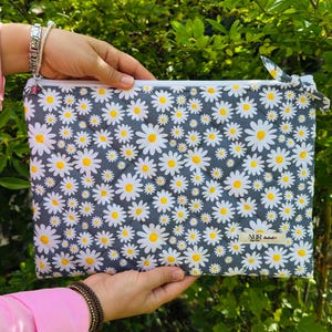 Könnte beinhalten: Eine graue, gesteppte Tasche mit weißem Reißverschluss und Gänseblümchenmuster. Die Gänseblümchen haben weiße Blütenblätter und gelbe Zentren. Die Tasche wird vor einem grünen, belaubten Hintergrund gehalten.