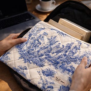 Könnte beinhalten: Eine gesteppte Laptoptasche mit blau-weißem Toile-de-Jouy-Muster wird in einem schwarzen Rucksack offen gehalten. Die Hülle zeigt eine detaillierte pastorale Szene. Ein Laptop und eine Tasse Kaffee sind im Hintergrund.