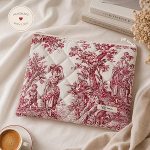 Peut inclure: Une pochette zippée matelassée rectangulaire avec un motif toile de jouy rouge et blanc représentant des personnages dans un paysage. Le tissu présente une texture piquée en losanges et une étiquette rectangulaire NUR Handmade.