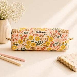 Könnte beinhalten: Ein rechteckiges Stifteetui aus Stoff mit einem Blumenmuster in Gelb-, Orange-, Rosa- und Grüntönen. Ein kleines beiges Stoffetikett auf der Vorderseite zeigt den Text NUR Design Hand-made mit einem kleinen roten Herzsymbol.