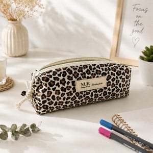 Puede incluir: Un estuche rectangular con estampado de leopardo en marrón y negro. El estuche tiene una cremallera de color crema y una etiqueta de tela blanca en la parte frontal con el texto NUR Design Kunst Handmade y un pequeño corazón rojo.