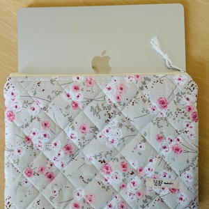 Könnte beinhalten: Eine gesteppte Laptoptasche mit einem Blumenmuster in Rosa- und Weißtönen auf beigefarbenem Hintergrund. Die Tasche hat einen Reißverschluss und einen kleinen weißen Quasten-Zug. Der Markenname "NUR" ist auf einem Etikett sichtbar, das an der Tasche befestigt ist.