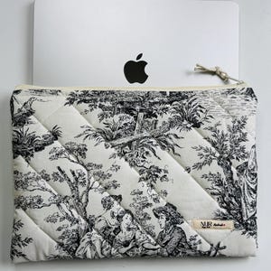 Può includere: Custodia per laptop trapuntata con un motivo toile de Jouy in bianco e nero raffigurante una scena pastorale. La custodia ha una cerniera color crema e una piccola etichetta. Un laptop argentato con il logo di una mela nera è parzialmente visibile.