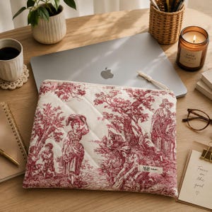 Op de afbeelding: Een rechthoekige gewatteerde laptophoes met een rood-wit toile de jouy-patroon met pastorale scènes met figuren en bomen. De hoes heeft een witte ritssluiting en een klein rechthoekig stoffen label met de tekst NUR.