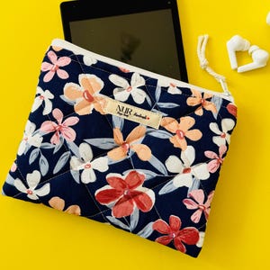 Könnte beinhalten: Eine blaue, gesteppte Reißverschlusstasche mit einem Blumendruck mit rosa, weißen und roten Blumen. Die Tasche hat einen weißen Reißverschluss und ein weißes Etikett mit dem Text "NUR Handmade". Die Tasche liegt auf einer gelben Oberfläche mit einem Tablet und weißen Ohrhörern.