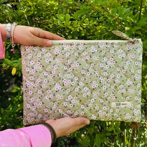 Pode incluir: Uma bolsa acolchoada retangular com um padrão floral de flores rosa sobre um fundo verde claro. A bolsa tem um fecho de correr e uma pequena presilha de tecido. A etiqueta "NUR" é visível no canto inferior direito.