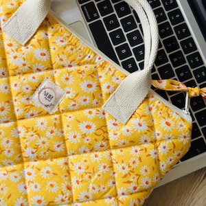 Könnte beinhalten: Gelb gesteppte Laptoptasche mit Gänseblümchen-Blumenmuster. Die Tasche hat einen cremefarbenen Griff und einen Reißverschluss. Ein kleines Etikett mit dem Text "NUR Design Book" ist auf der Vorderseite aufgenäht. Die Tasche liegt auf einer Laptop-Tastatur.