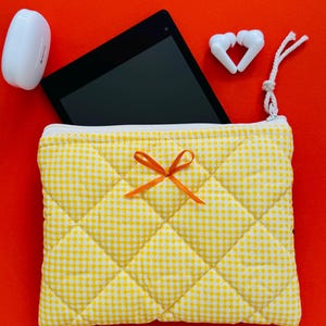 Op de afbeelding: Een gele en witte geruite, gewatteerde tas met een oranje strik. De tas ligt op een rood oppervlak met een witte tablet en witte oordopjes.