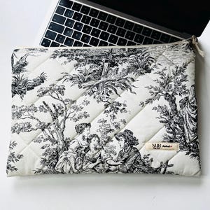 May include: Cream-colored quilted laptop sleeve with a black and white toile design featuring figures in a landscape. The sleeve has a zipper closure and a small tag with text. A laptop is partially visible.