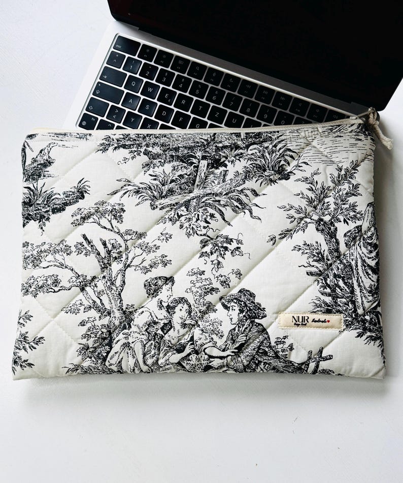 May include: Cream-colored quilted laptop sleeve with a black and white toile design featuring figures in a landscape. The sleeve has a zipper closure and a small tag with text. A laptop is partially visible.