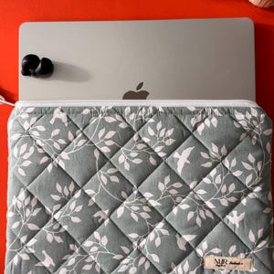 Könnte beinhalten: Eine graue, gesteppte Laptoptasche mit einem weißen Blumenmuster und einem kleinen weißen Etikett mit dem Text "NJR Handmade".