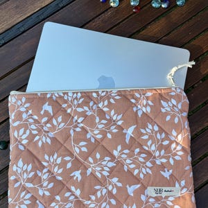 Könnte beinhalten: Eine gepolsterte Laptop-Hülle mit einem pfirsichfarbenen Hintergrund und einem weißen Blumen- und Vogelmuster. Die Hülle hat einen Reißverschluss und ein kleines Etikett mit der Aufschrift "NUR Handmade".
