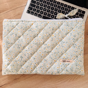 Puede incluir: Funda acolchada para portátil con un estampado floral de margaritas blancas y amarillas sobre un fondo verde claro. La funda tiene una cremallera blanca y una pequeña etiqueta de tela. Un teclado de portátil es visible en el fondo.