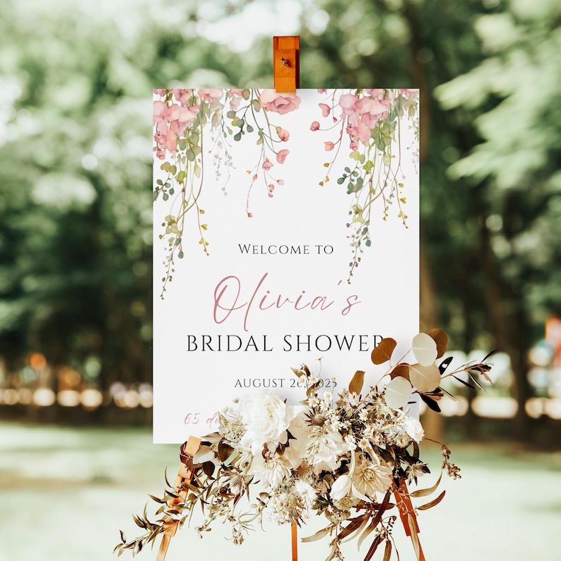 May include: A white sign with the text "Welcome to Olivia's Bridal Shower" in a script font, accented with pink floral illustrations. The sign is propped up on a wooden easel and decorated with a floral arrangement.