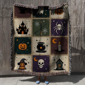 May include: A beige Halloween-themed throw blanket with a grid pattern of spooky illustrations. The squares feature images of a black cat, ghost, pumpkin, haunted house, spider, witch, cauldron, skull, and candle. The blanket has a fringed edge.
