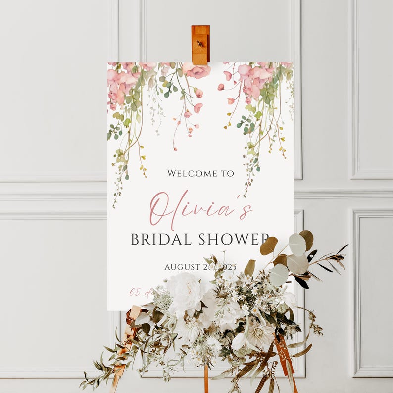May include: A white sign with floral accents and the text "Welcome to Olivia's Bridal Shower" in a script font. The sign is decorated with pink flowers and greenery. The date August 20, 2023 is also displayed.
