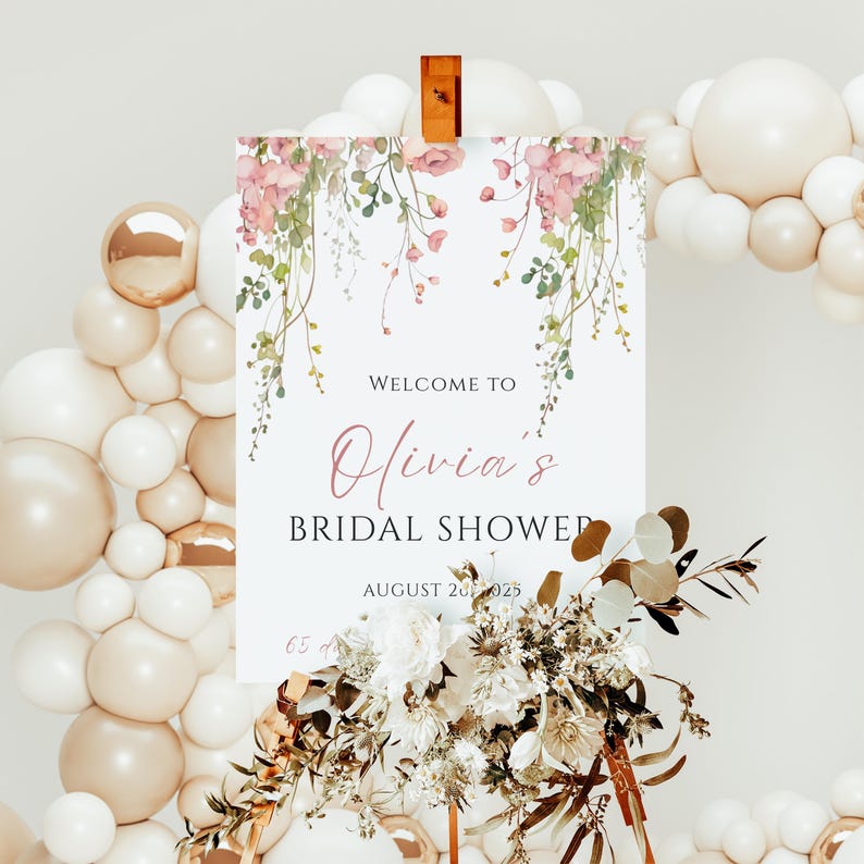 May include: A white sign with floral accents and the text "Welcome to Olivia's Bridal Shower" is displayed. The sign is decorated with pink flowers and greenery. A balloon arch and floral arrangement surround the sign.