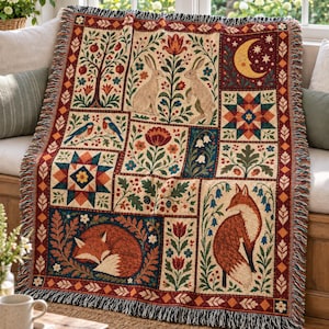 May include: A woven throw blanket featuring a multi-panel design with illustrations of rabbits, a fox, birds, a crescent moon, stars, and floral motifs in red, blue, green, and cream. The blanket has a fringed edge and a decorative geometric border.