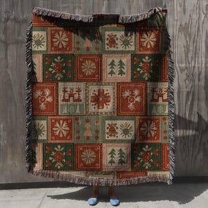 Puede incluir: Manta tejida con diseño de patchwork. Los cuadrados son rojos, verdes y beige, con copos de nieve, árboles y motivos florales. La manta tiene un borde con flecos. Dimensiones no visibles.