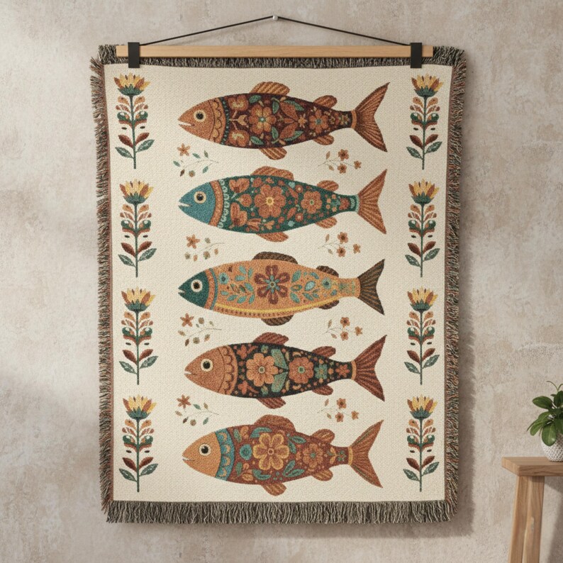 May include: A cream-colored woven throw blanket featuring a repeating pattern of colorful fish with floral designs. The fish are in shades of brown, orange, and teal. The blanket is hung on a wooden rod and has a fringed edge.