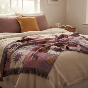 May include: A woven throw blanket with a celestial design featuring suns and moons in shades of pink, purple, and gold. The blanket is draped over a bed with pillows in shades of pink and gold. The blanket has a fringed edge.