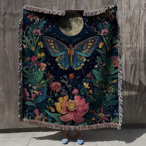 May include: A dark blue woven throw blanket featuring a large butterfly with blue and gold wings, surrounded by colourful flowers and foliage. A full moon is visible in the background. The blanket has a fringed edge.